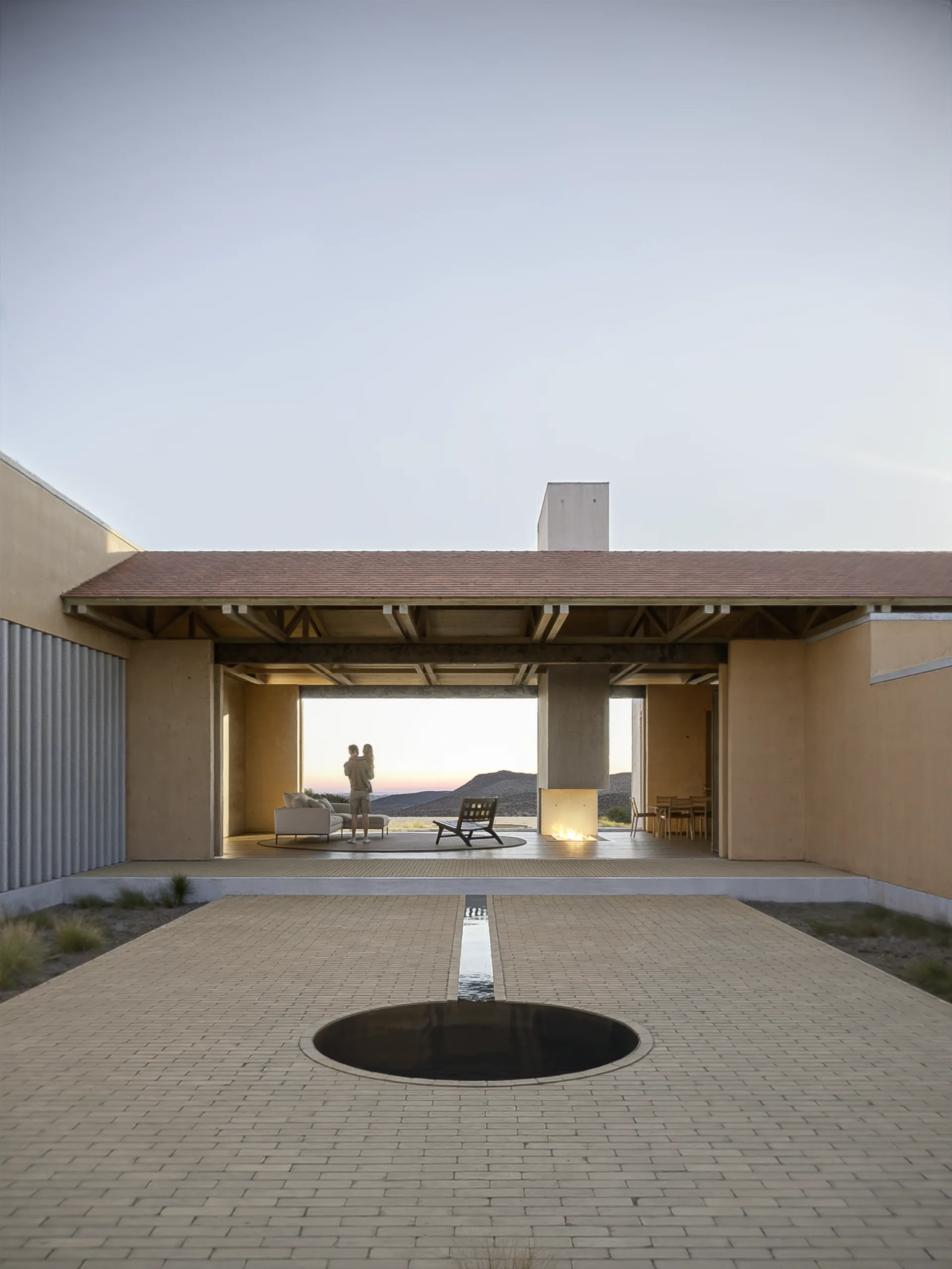 Render del patio central de la casa muestra de Aldea X al atardecer, con espejo de agua y chimenea encendida